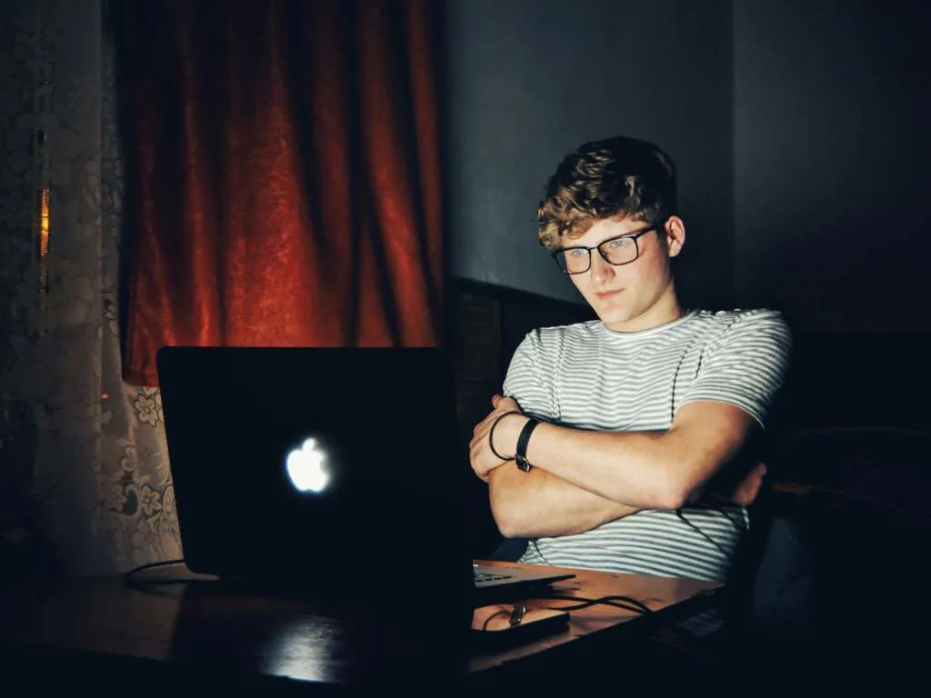 LGBTQ young man in glasses sitting alone in a dark room, illuminated by a laptop screen, appearing thoughtful or emotionally withdrawn — symbolizing late-night reflection or silent struggle.