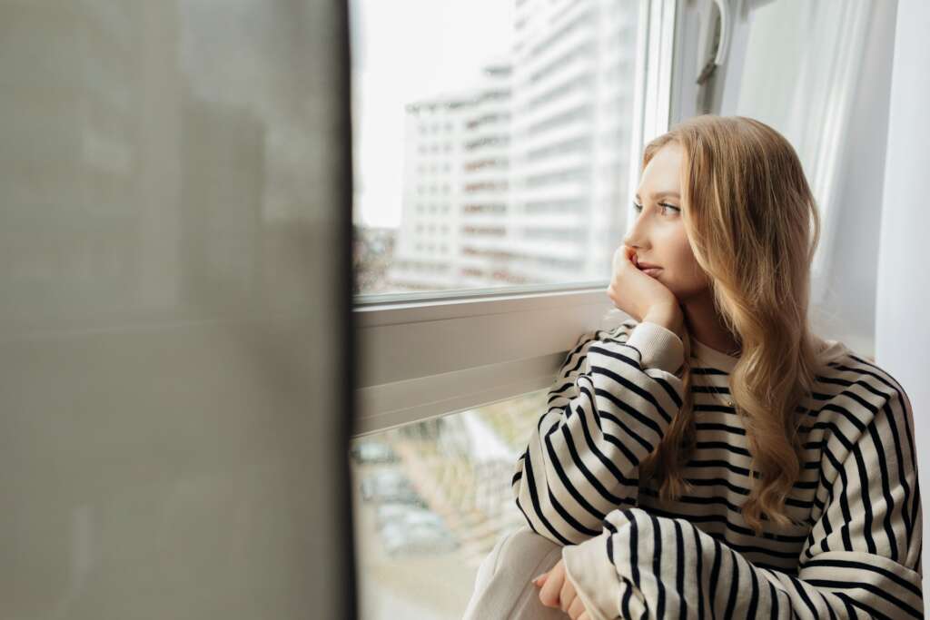 Woman sitting by a window in soft natural light, quietly reflecting and holding unspoken emotions.